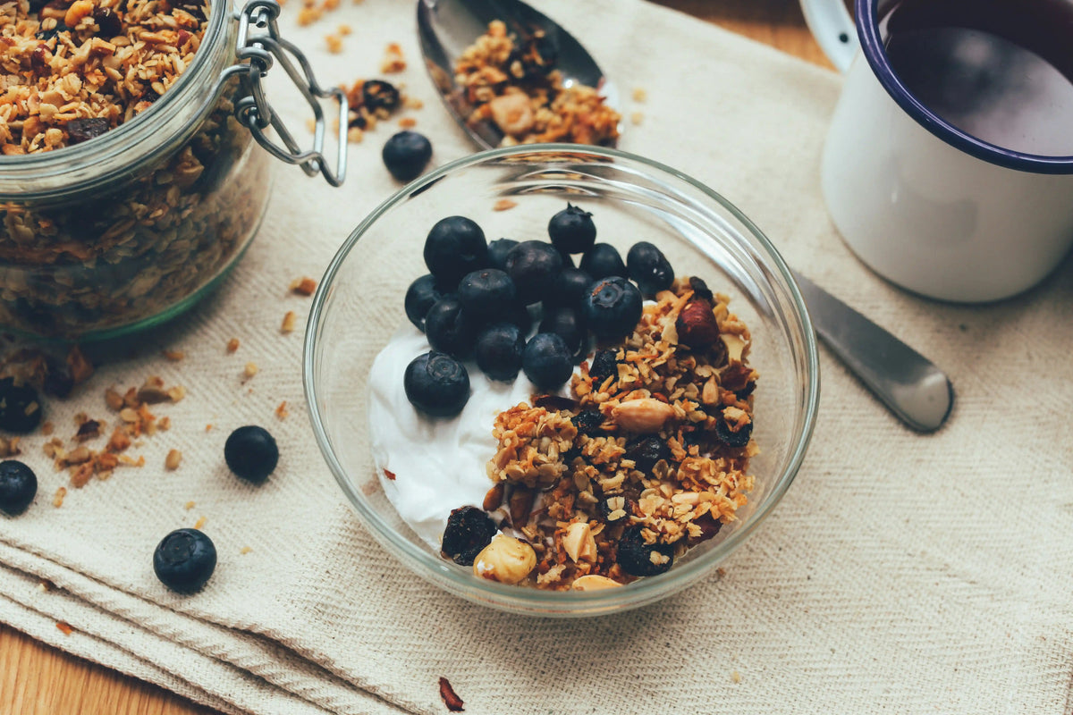 Glasskål med yoghurt, blåbär och granola med nötter och torkad frukt på en ljus linneduk.